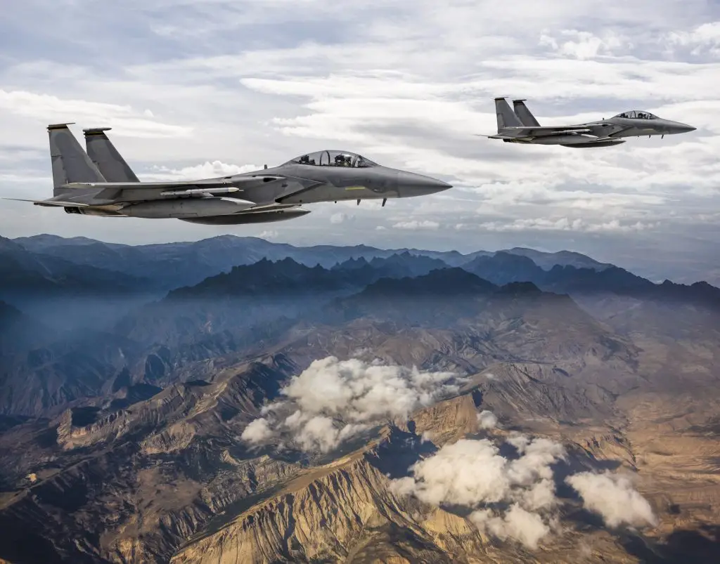 Two fighter jets fly in formation over a landscape of rugged mountains and clouds under a partly cloudy sky.