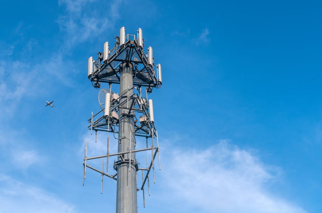 Une tour de téléphonie mobile se dresse dans un ciel bleu parsemé de nuages. Un petit avion est visible au loin, à gauche de la tour.