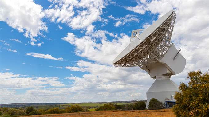 Large radio telescope dish facing a blue sky with scattered clouds, situated in a field with distant scenery.