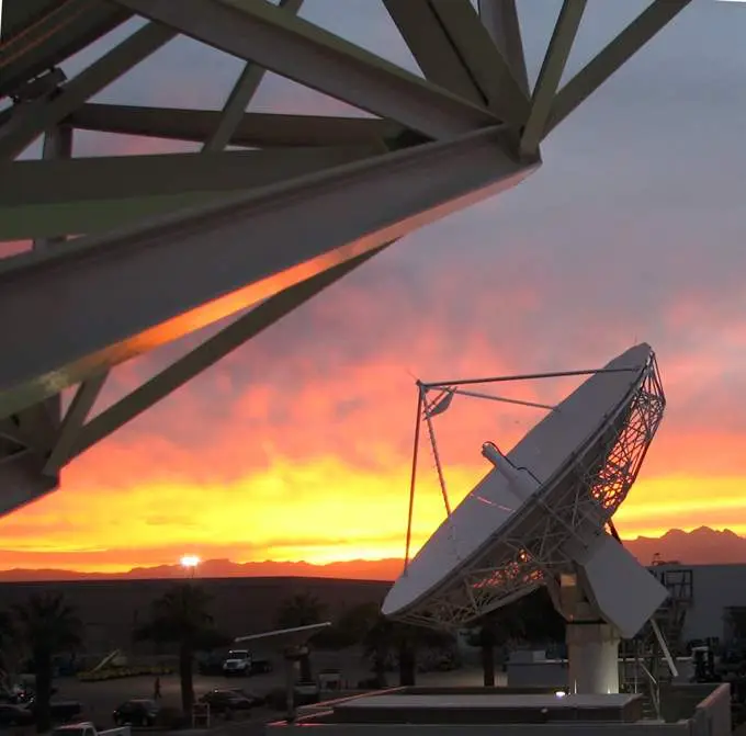 A satellite dish at sunset with vibrant orange and pink clouds in the background and a geometric metal structure in the foreground.