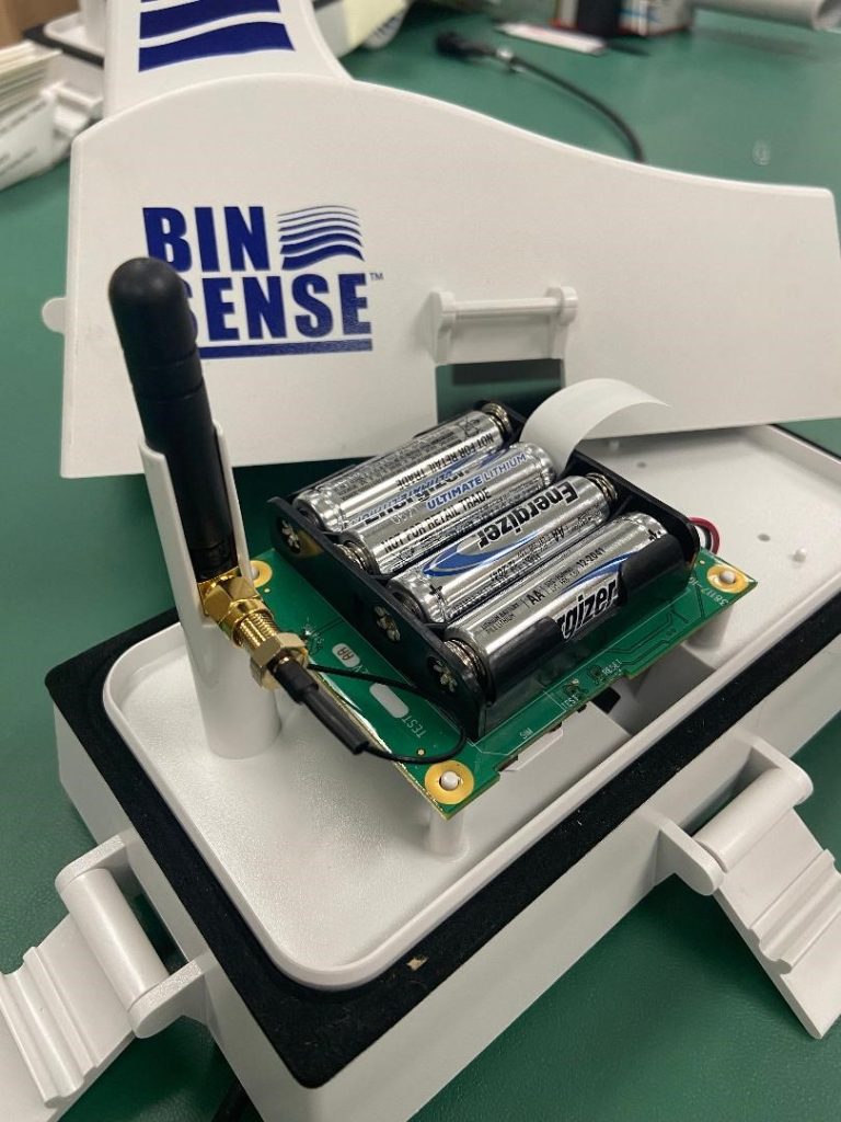 Electronic device with four visible batteries and a connected antenna, branded "BIN SENSE", on a green worktable.