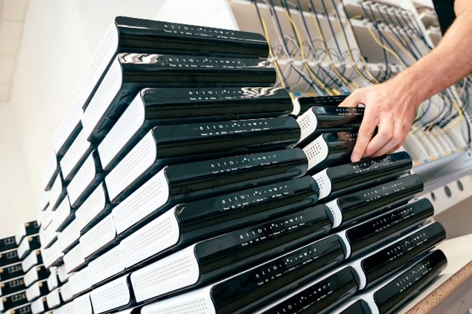 A person's hand arranging a stack of black laptops on shelves, each connected by a cable for charging or data synchronization.