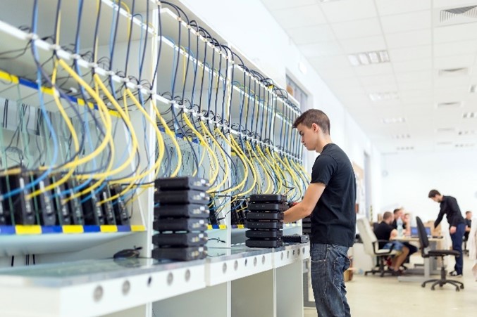 A man working in a server room, connecting cables to network devices with colleagues in the background.