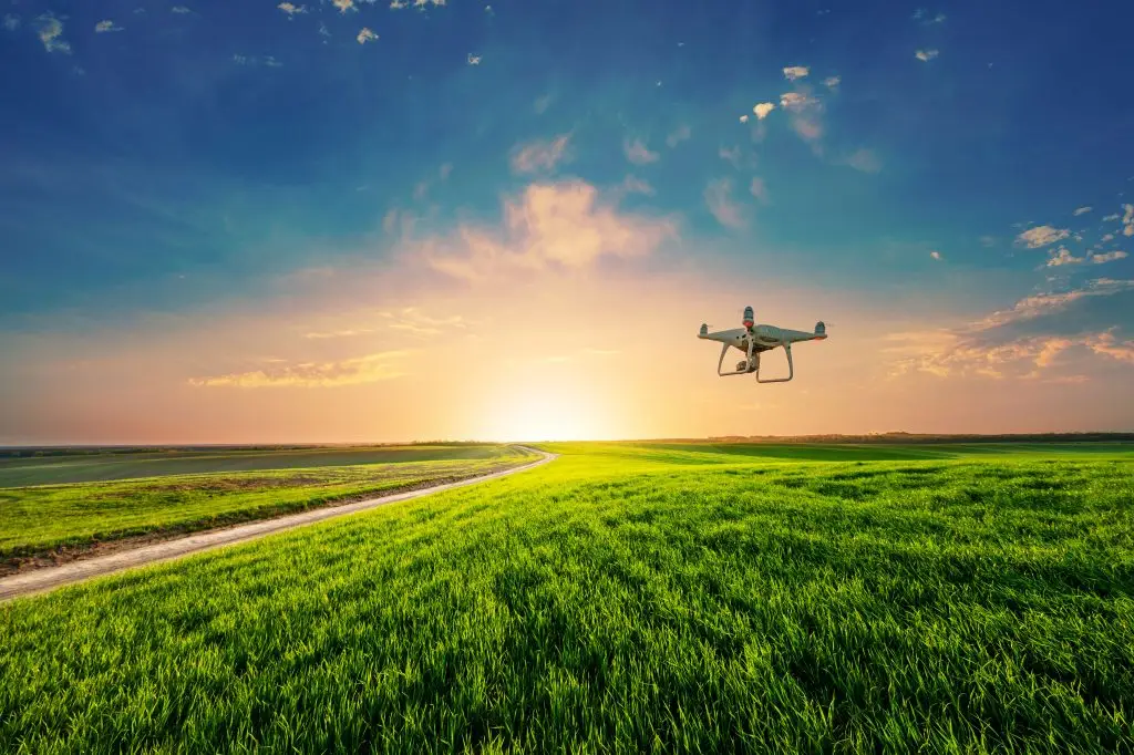 Un drone survole un champ verdoyant au coucher du soleil, avec un chemin sinueux menant à l'horizon sous un ciel partiellement nuageux.