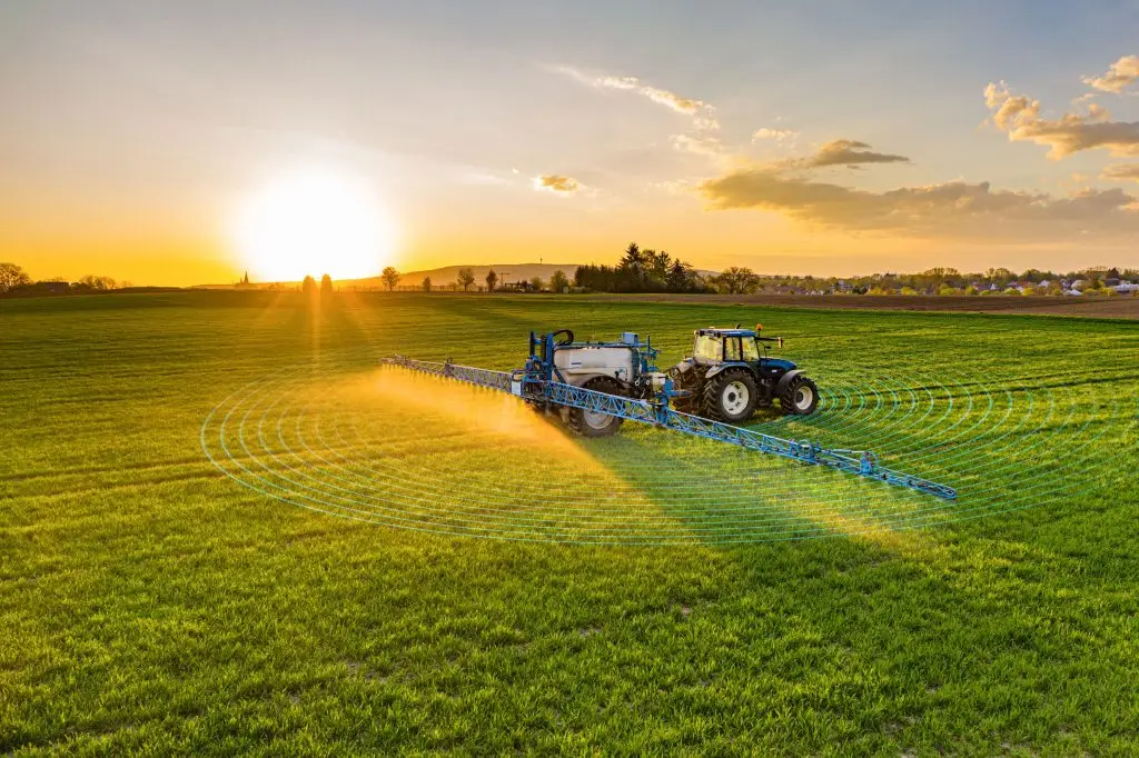 A tractor sprays a crop field with pesticide at sunset, illustrating agricultural automation and precision farming.