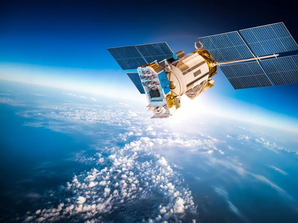 Un satellite équipé de panneaux solaires est en orbite au-dessus de l'atmosphère terrestre, avec une vue sur les nuages et le ciel bleu en dessous.
