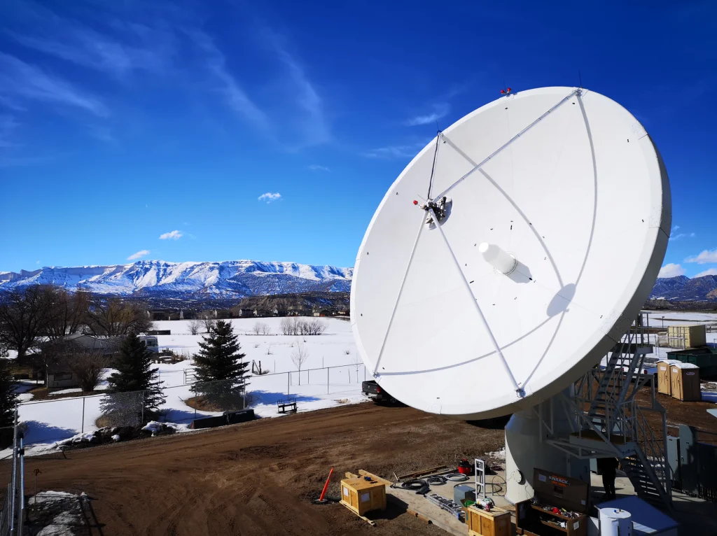 Large satellite dish in a snowy landscape with mountains in the background under a clear blue sky. Equipment and boxes are visible on the ground nearby.