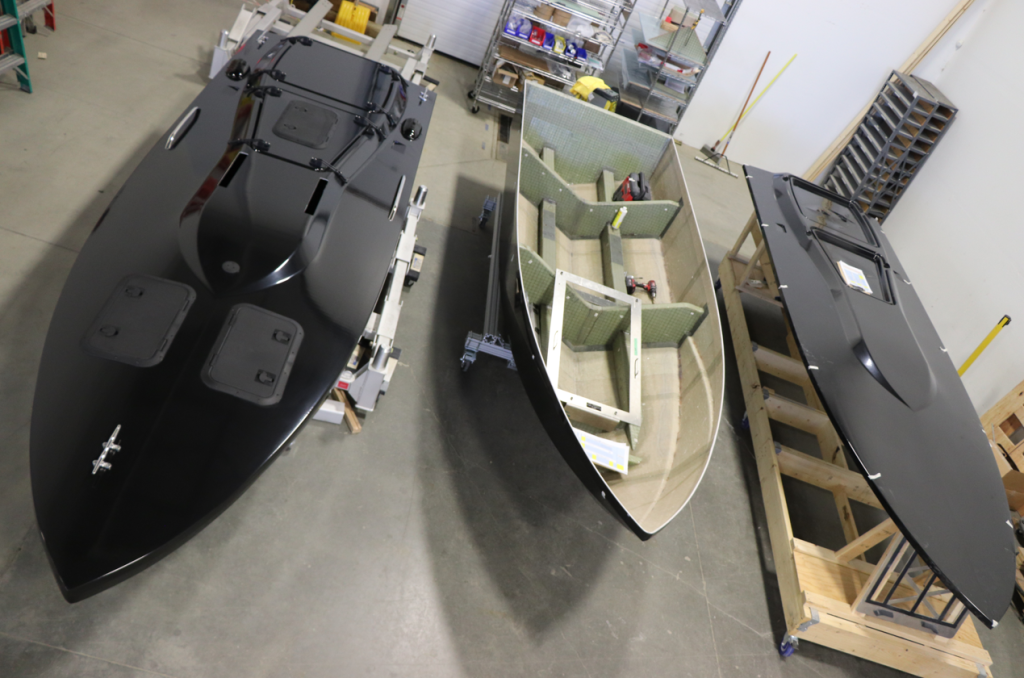 Three boats in a workshop: two black with sleek finishes, one partially assembled with visible framework. Shelves and tools are visible in the background.