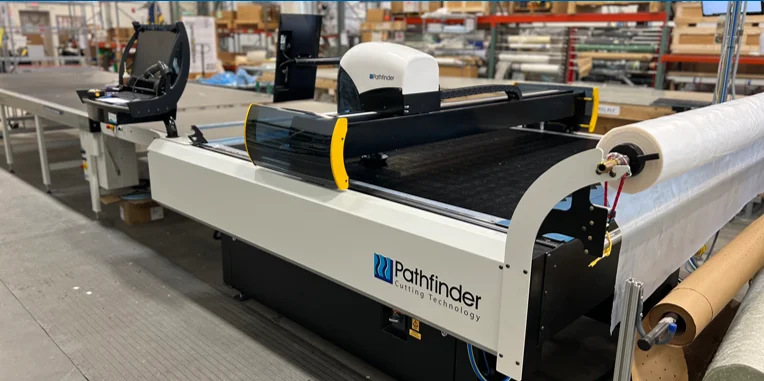 A cutting machine branded "Pathfinder Cutting Technology" in a factory setting. The machine has a roll of material and a control system.