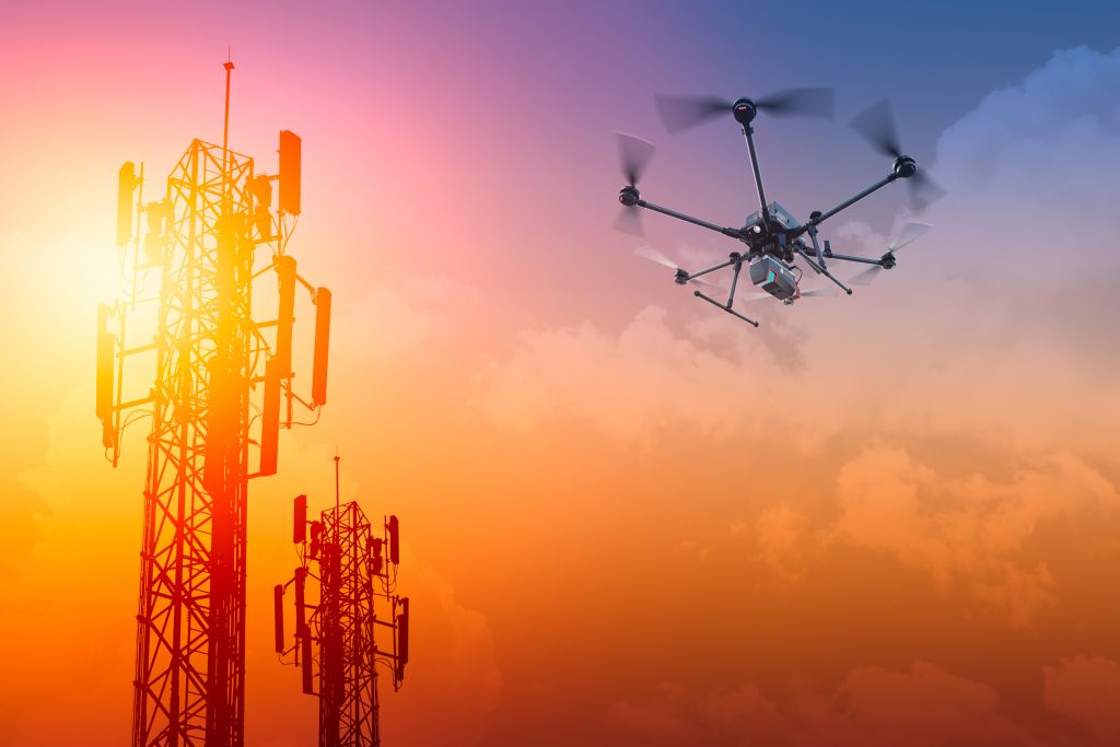 A drone with six propellers flies near two telecommunications towers during sunset, under a sky with orange and blue hues.