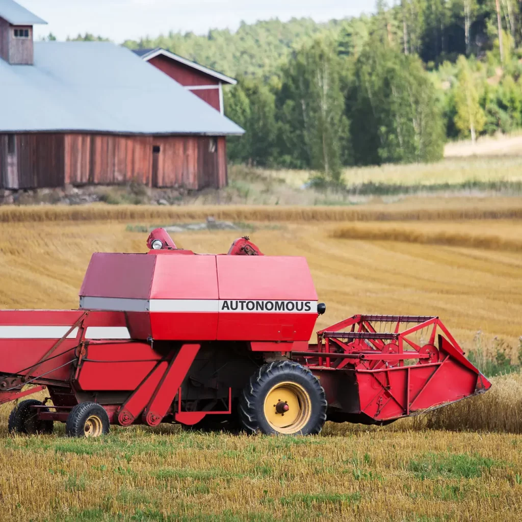 A red autonomous combine harvester operates in a wheat field next to a barn.