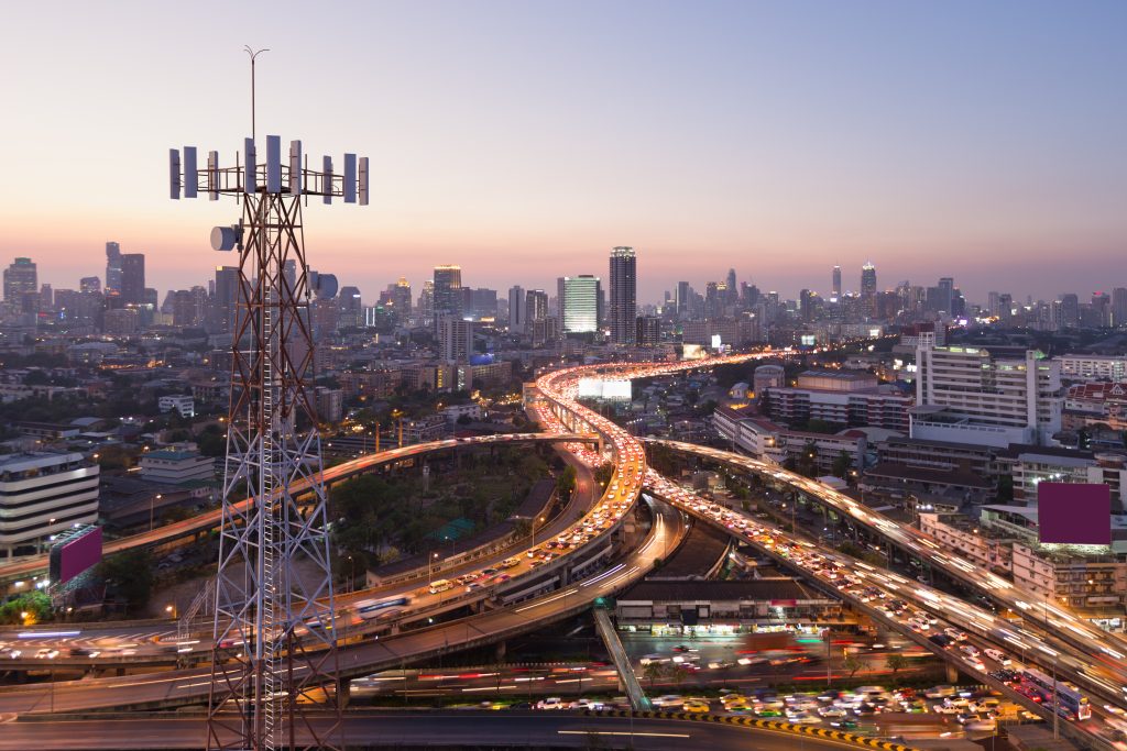 Aerial view of a bustling cityscape at dusk with busy highways, tall buildings, and a telecom tower in the foreground.