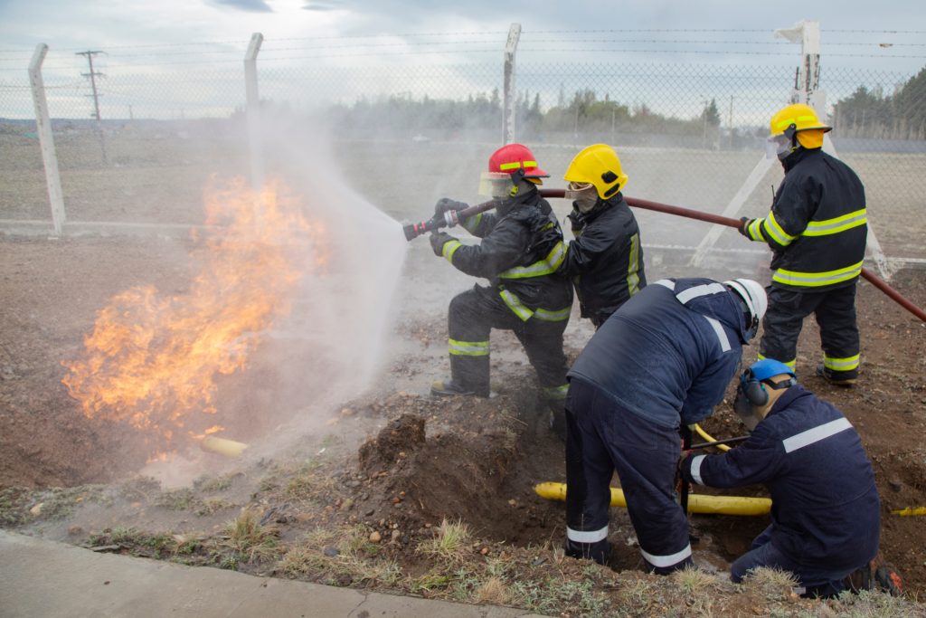 Four firefighters in protective gear extinguish a ground fire with hoses while two workers handle a yellow pipe, near a barbed wire fence.