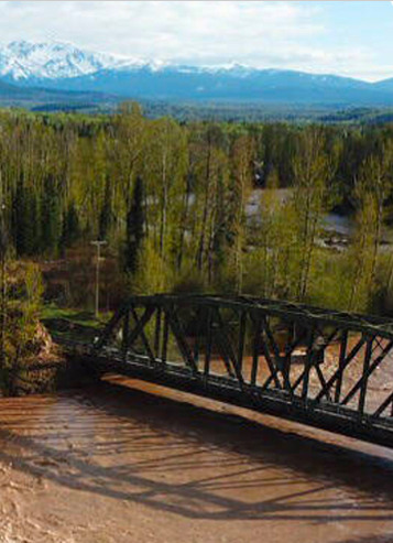 A metal bridge crosses a muddy river with a forest and snow-capped mountains visible in the background under a partly cloudy sky.