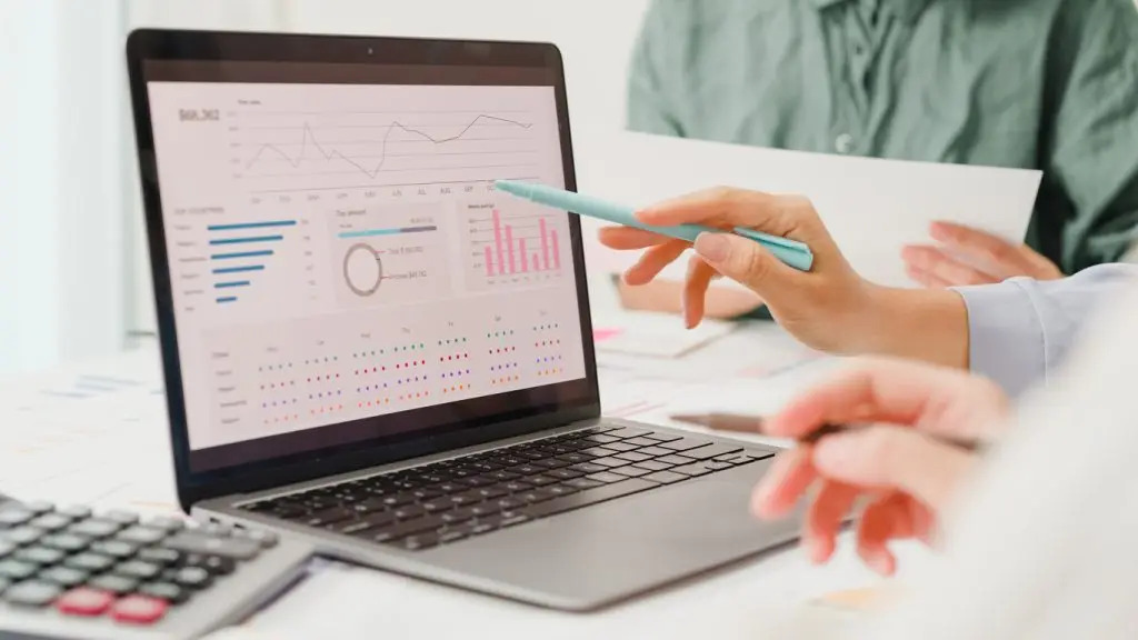 Two people review data visualizations and charts on a laptop screen, using a pen to point at information. A calculator and documents are on the desk.