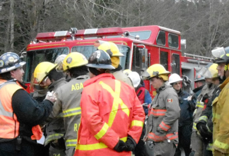 A group of firefighters in protective gear stand in front of a red fire truck, appearing to discuss their response at an emergency scene.