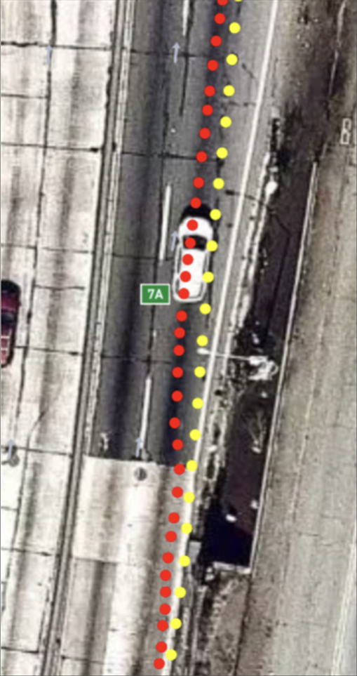 Aerial view of a white vehicle on a highway next to a solid line of red, yellow, and orange dots, with a green "7A" exit sign visible on the road.