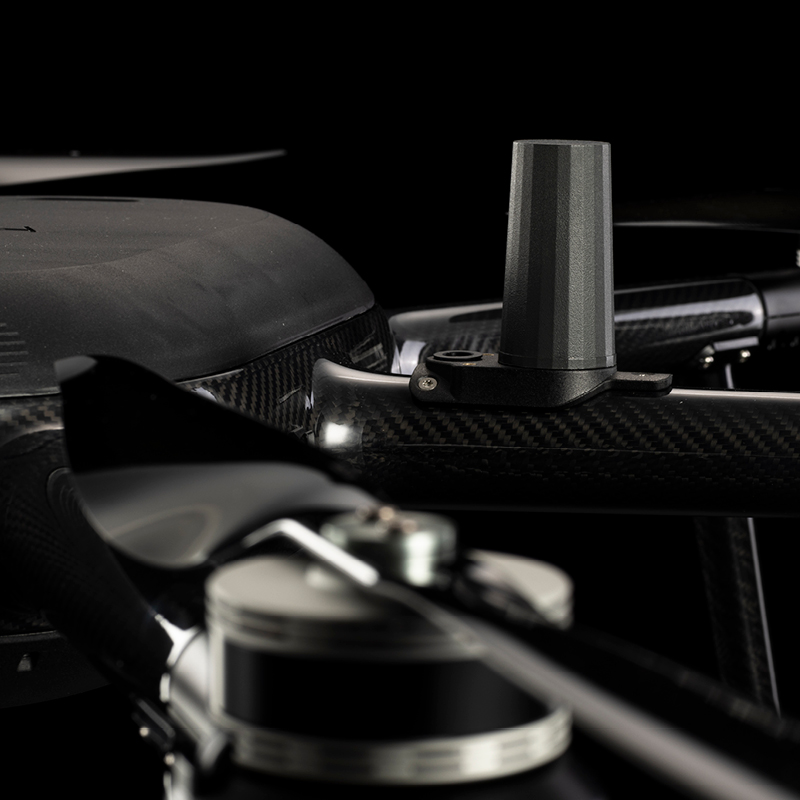 Close-up view of a drone's carbon fiber arm, showing a propeller, motor, and a small cylindrical sensor or antenna mounted on the arm against a dark background.