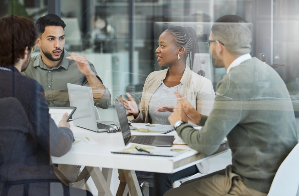Four people sit around a table with laptops and documents, engaged in discussion in a modern office setting.