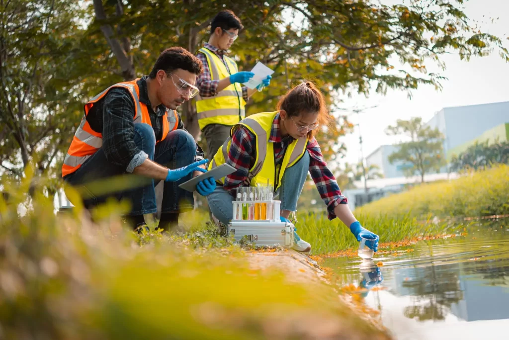 Three people wearing safety vests and gloves conduct water quality testing at the edge of a pond, using various scientific equipments and taking notes. Trees and a building are in the background.