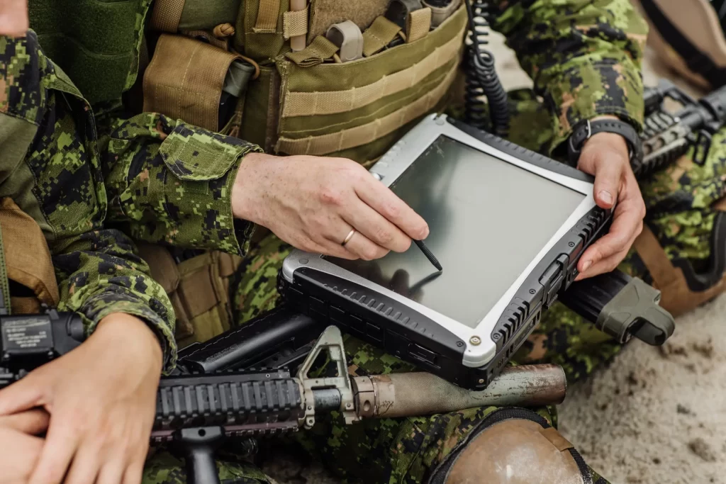 Two military personnel in camouflage uniforms use a rugged tablet with a stylus. A rifle is seen in the foreground.