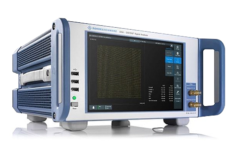 A digital signal analyzer with a screen displaying data, multiple input/output ports, and control buttons on the front panel. The device has a handle on the side and is branded Rohde & Schwarz.
