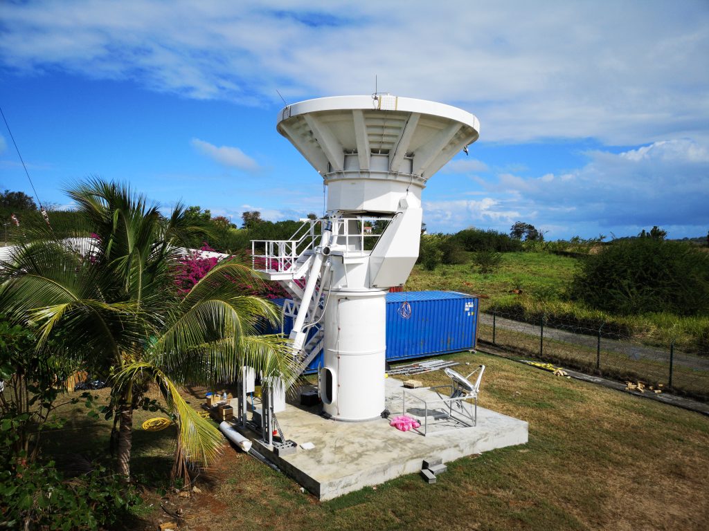 An outdoor radar installation with a large white dish and supporting structure, surrounded by greenery, palm trees, and a blue shipping container. The sky is partly cloudy.