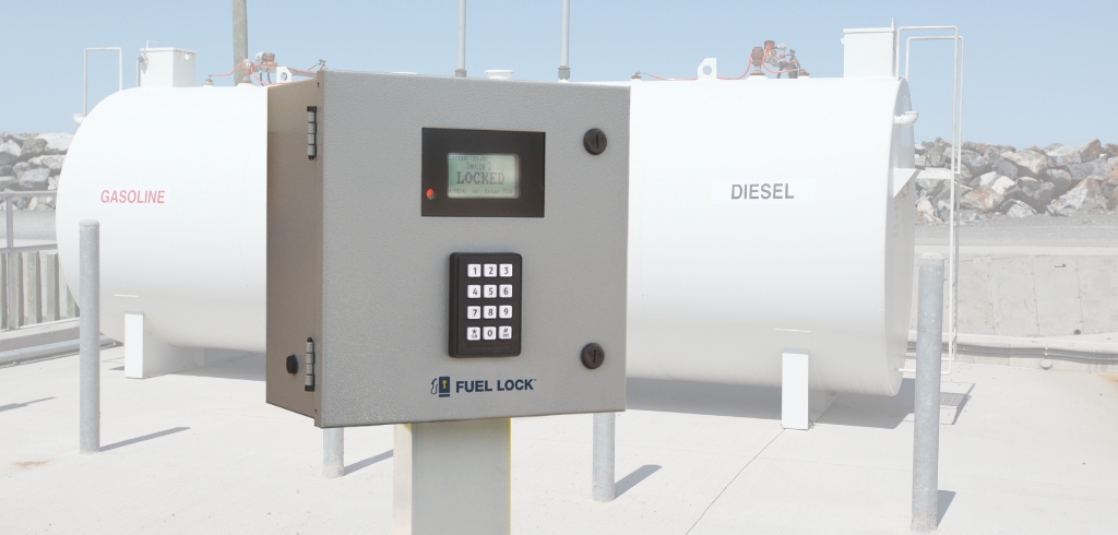 Close-up of a Fuel Lock control panel with a keypad and display screen, installed outdoors in front of two large fuel storage tanks labeled "Gasoline" and "Diesel".