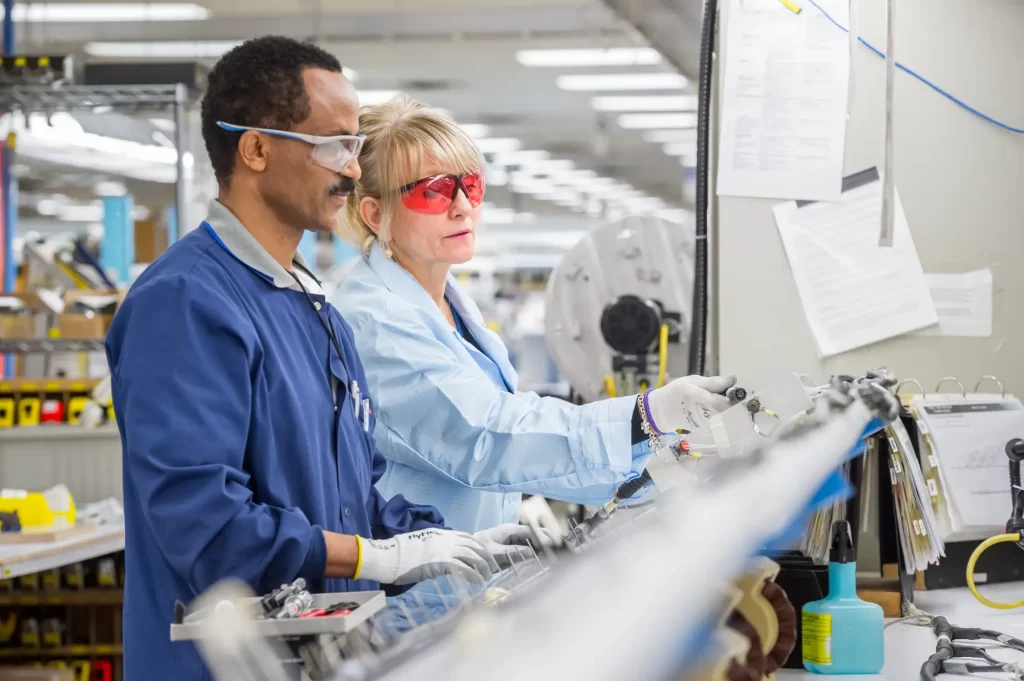 Two factory workers in safety gear operate machinery in a well-lit manufacturing setting. They both wear protective gloves, glasses, and blue lab coats.