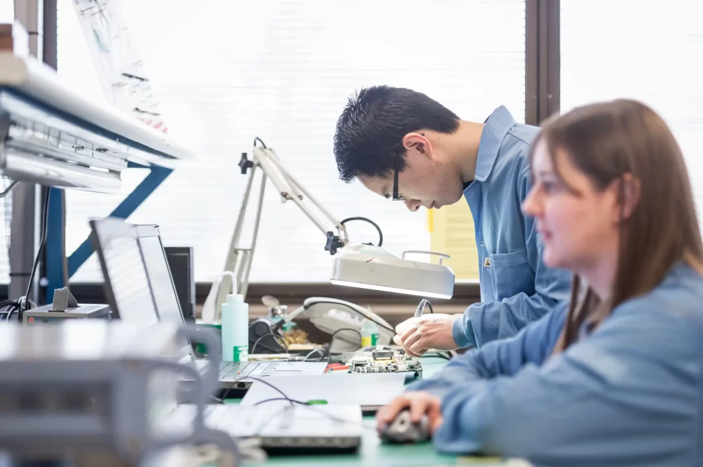 Two people work at a lab bench with computers and electronic equipment, one is focused on soldering a circuit board while the other works on a computer.