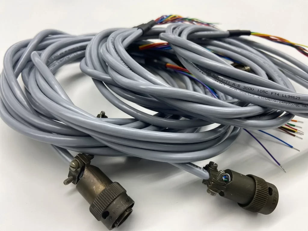 Coiled grey electrical cables with connectors on the ends and various wires exposed on the other side, placed on a white surface.