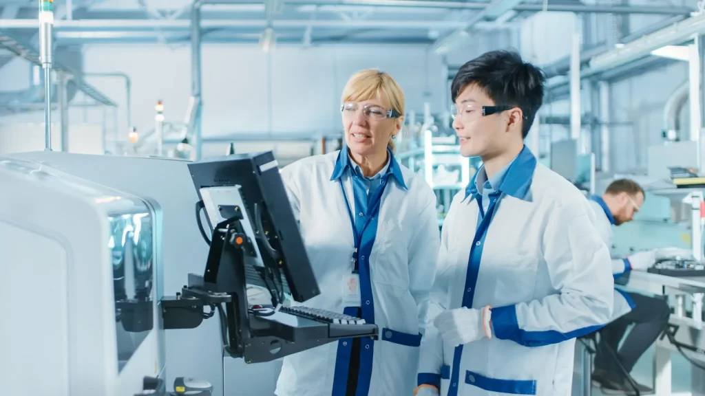 Two lab technicians in lab coats and safety glasses work at a control panel. They appear to be discussing something on the screen. The lab is equipped with various machinery and instruments.