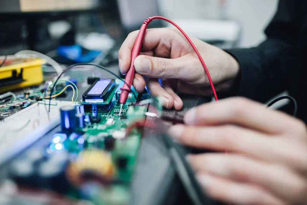 Close-up of a person using a red tool to test an electronic circuit board with various components and wires.