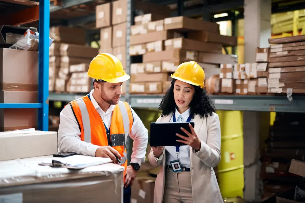 Two people wearing safety gear and hard hats work in a warehouse. One holds a clipboard, and the other holds a tablet, surrounded by stacks of boxes.