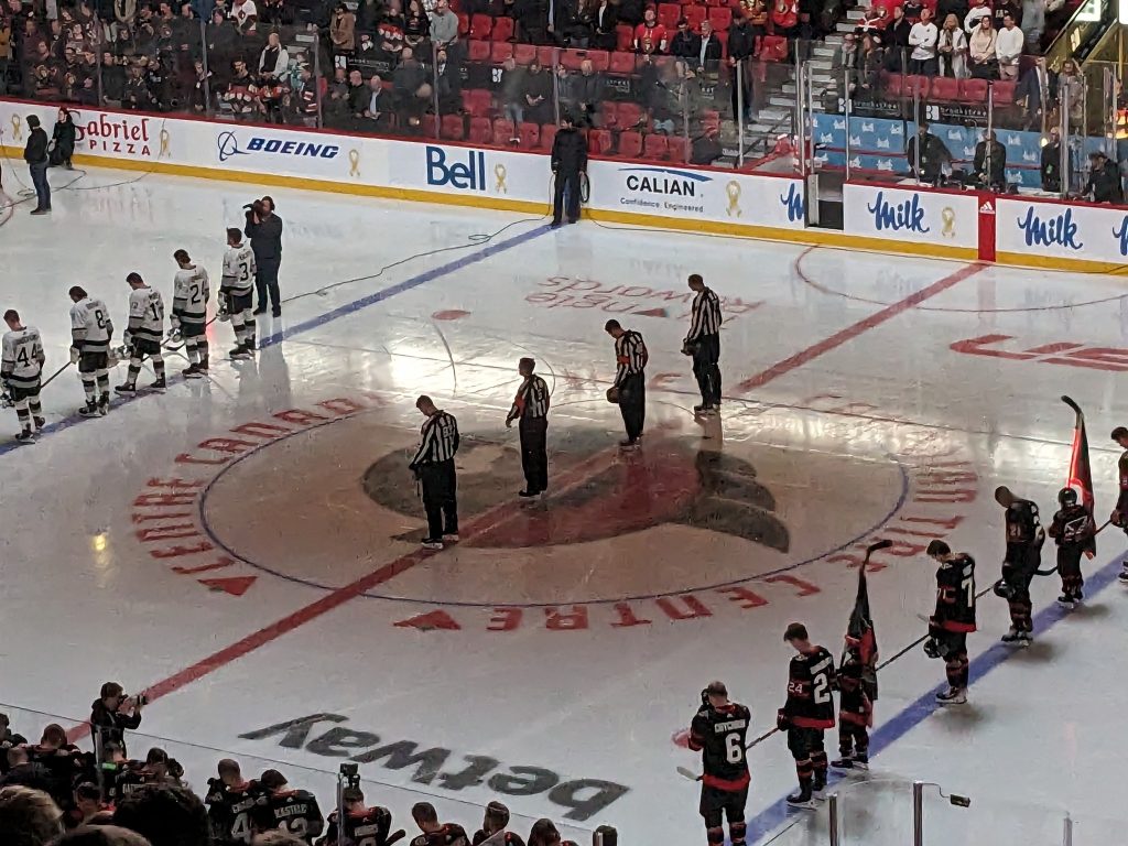 Hockey teams line up for pre-game ceremonies on an ice rink, with referees standing at center ice. Spectators are seated in the background.