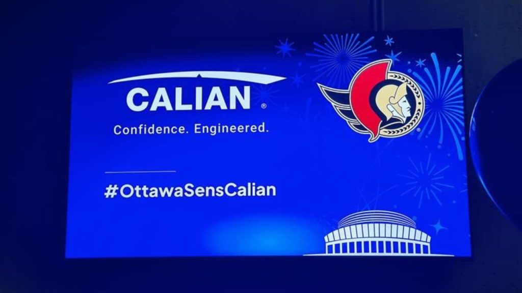 Un panneau bleu affiche le logo Calian, le logo des Sénateurs d'Ottawa et le mot-clic #OttawaSensCalian avec des feux d'artifice et un bâtiment illustré en arrière-plan.