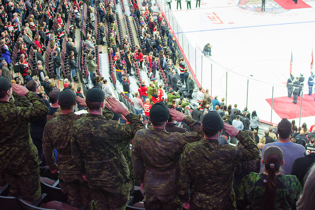 Soldiers in camo salute during a hockey game as the audience stands.