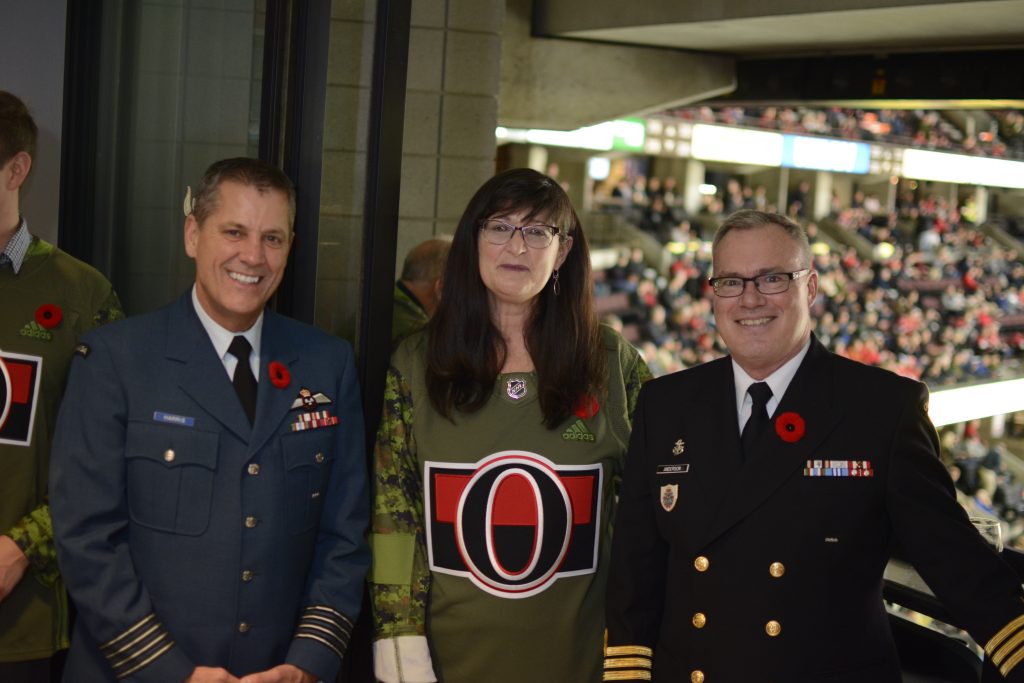 Trois personnes posent pour une photo, deux en uniforme militaire et une en maillot de sport, toutes portant des coquelicots rouges. L'arrière-plan montre un stade avec des spectateurs debout.