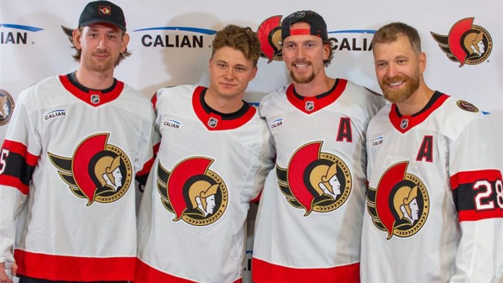 Four ice hockey players wearing white jerseys with a red, black, and gold logo stand side by side and smile for a photo.