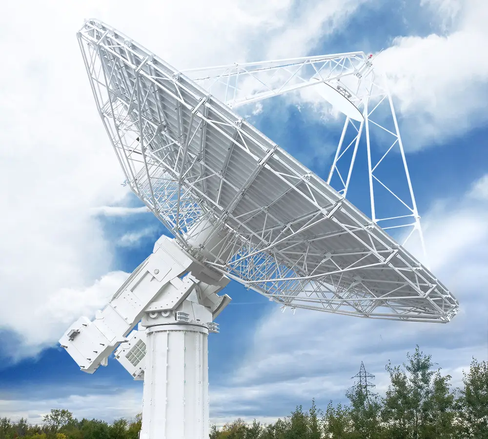 A large white satellite dish antenna is pointed towards the sky, set against a background of clouds and blue sky, with some trees at the base.