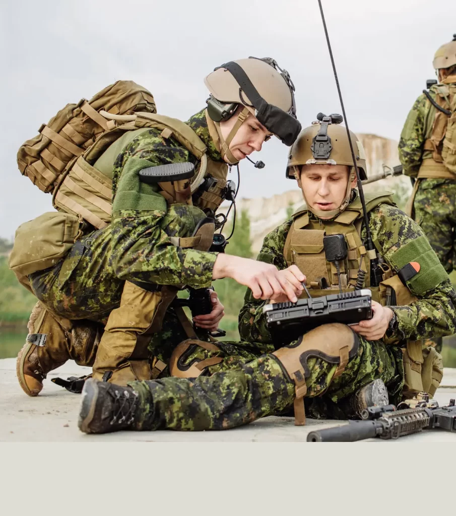 Two soldiers in camouflage uniforms, kneeling and sitting, examine a handheld device outdoors. One holds a rifle, the other adjusts the device. A third soldier stands in the background.