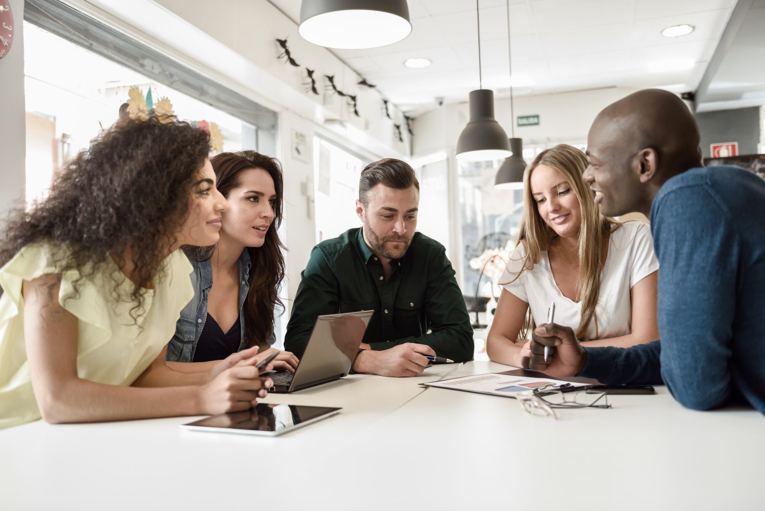 A diverse group of people sit around a table with laptops and tablets, having a discussion in a bright room.