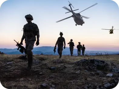 Silhouetted soldiers walk across a field at sunset, with helicopters flying overhead.