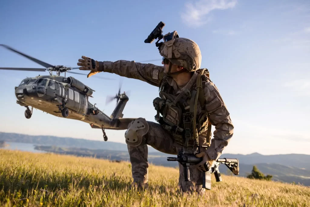 Soldier in gear crouches in a field, gesturing towards a military helicopter taking off.