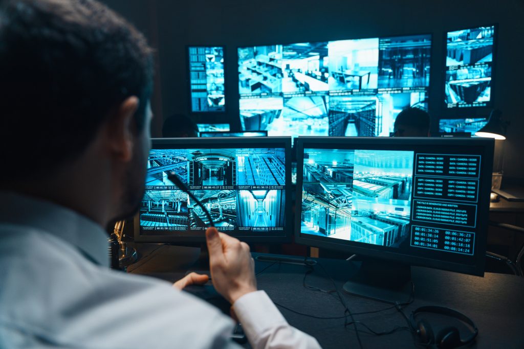A person monitors security footage on multiple computer screens in a control room filled with technology equipment and large display monitors.