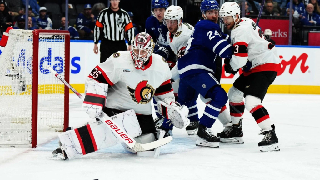 A hockey game in action with players in blue and white jerseys colliding with players in white and red jerseys near the goal, and a goalie in white and red gear guarding the net.