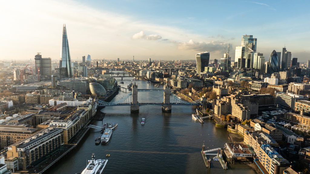 Aerial view of London featuring the River Thames, Tower Bridge, and numerous skyscrapers, including the Shard, under a partly cloudy sky.