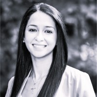 A black and white photo of a smiling person with long straight hair, wearing a light blouse with a cross necklace, in front of a blurred outdoor background.