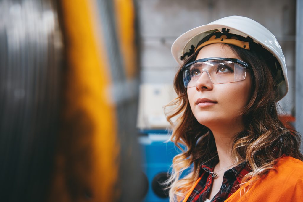 A woman in a hard hat and safety glasses looks up, standing in an industrial setting with blurred machinery in the foreground.