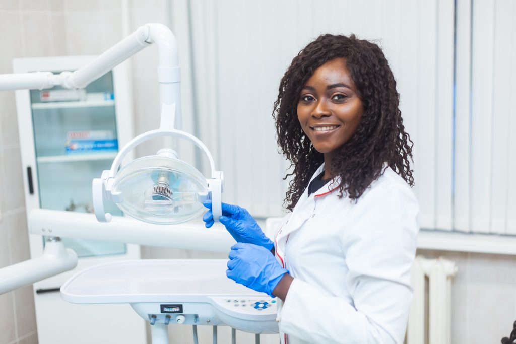 A dentist wearing a white coat and blue gloves stands next to a dental chair and equipment, smiling at the camera in a clinic setting.